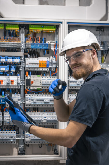 Electrical technician working in a switchboard with fuses, uses a tablet. Happy bearded technician in protective helmet with digital tablet.